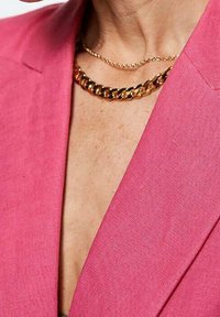 Blazer in lino fucsia con scollatura a V profonda, abbinato a collane d'oro a strati: una catena delicata e una catena a maglie grosse.