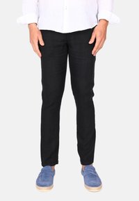 Schwarze, schmal geschnittene Jeanshose mit subtiler Textur. Kombiniert mit blauen Slippern mit Jutesohle.