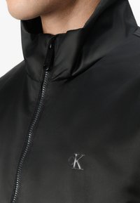 Zwarte zip-up jas met hoge kraag, gemaakt van een glad materiaal, voorzien van een subtiele logo aan de linkerkant en een elegante rits.