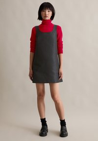 Γκρι υφασμάτινο φόρεμα με τετράγωνη λαιμόκοψη, φορεμένο πάνω από μια φωτεινή κόκκινη μπλούζα με ψηλό λαιμό. Το μοντέλο φοράει μαύρες κάλτσες και χοντρές μαύρες παπούτσια.