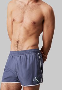 Pantaloni da bagno blu profondo con dettagli bianchi, vita elastica e logo cK verde chiaro. Realizzati in tessuto leggero e morbido.