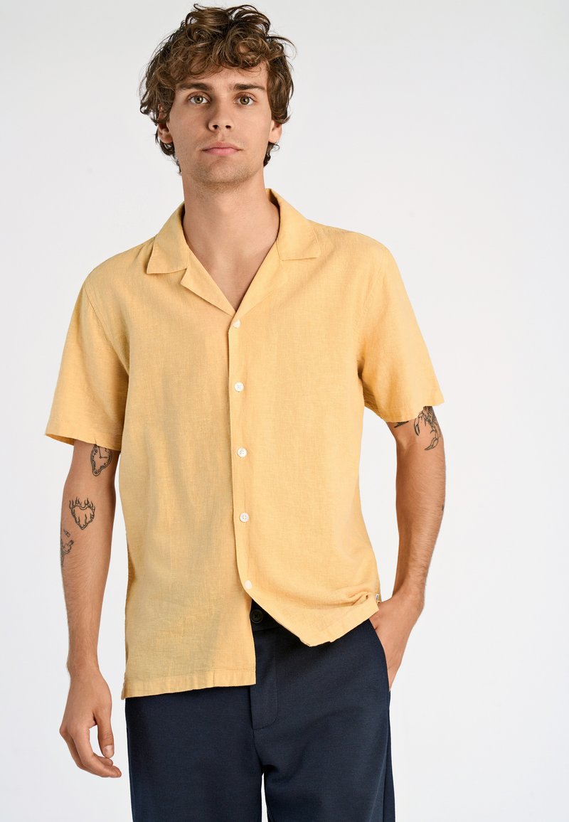 Giovane uomo con capelli ricci che indossa una camicia gialla a maniche corte e pantaloni scuri, in piedi con una mano in tasca.