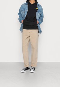 Hellblaue Jeansjacke, schwarze Kapuzenjacke mit Logo, beige zugespitzte Hose und schwarze Sneaker mit weißen Sohlen. Einfaches, lässiges Outfit.