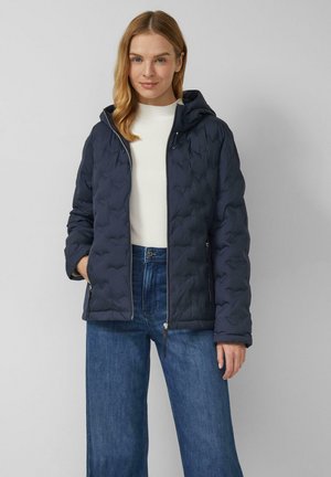 Femme portant une veste matelassée à capuche bleu marine par-dessus un haut blanc et un jean bleu, debout devant un fond gris clair uni.