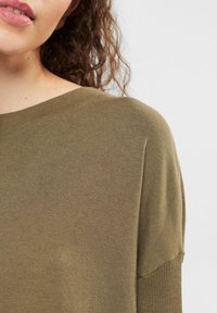 Pull en tricot vert olive avec un col rond. Dispose d'une texture lisse et de manches courtes à emmanchure descendante.