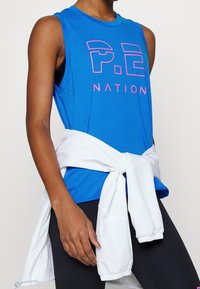 Camiseta atlética sin mangas de color azul con el logo rosa de "P.E NATION". Chaqueta blanca atada a la cintura. Leggings negros de textura suave.