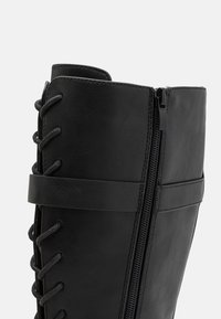 Bottine en cuir noir avec un haut tige, fermeture éclair latérale et design à lacets à l'avant avec des lacets gris, complétant une texture lisse.