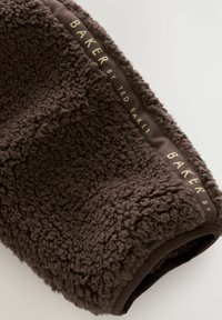 Coperta in pile marrone con una superficie morbida e testurizzata. Ha un bordo in tessuto scuro liscio e una scritta dorata che recita "BAKER di TED BAKER."