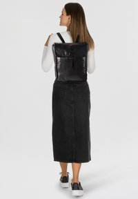 Mochila de cuero negro con solapa plegable, con cierre de correa y correas de hombro ajustables. El modelo lleva una falda de mezclilla negra y zapatos.