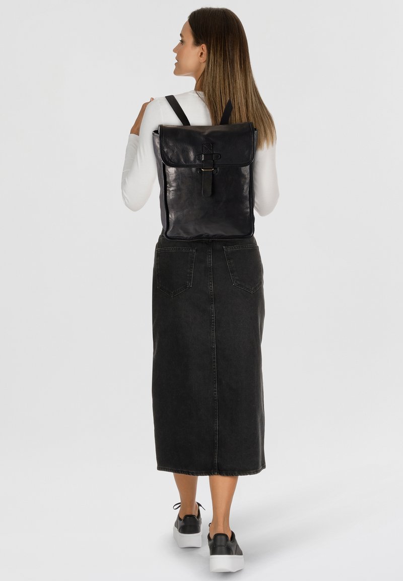 Mochila de cuero negro con solapa plegable, con cierre de correa y correas de hombro ajustables. El modelo lleva una falda de mezclilla negra y zapatos.