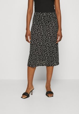 Femme portant une jupe midi noire à pois blancs, un haut noir sans manches et des sandales tissées noires à talons devant un fond uni.