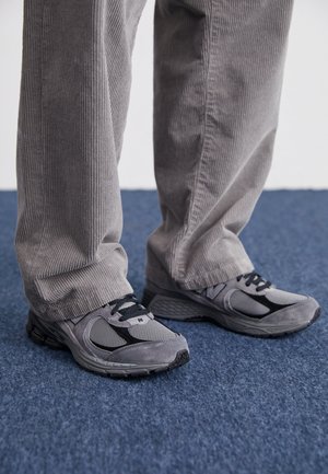 Ténis atléticos cinzentos com parte superior em malha e camurça, design com atacadores, sola texturizada, colocados sobre calças de veludo cotelê cinzentas num tapete azul.