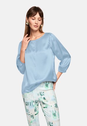 Light blue satin blouse with a boat neckline and 3/4 sleeves, paired with patterned trousers in green and white geometric designs.