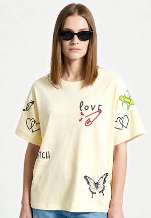 Junge Frau trägt schwarze Cat-Eye-Sonnenbrille und ein cremefarbenes T-Shirt mit Illustrationen von Liebe, Schmetterling, Herzen und Pflaster sowie blaue Jeans.