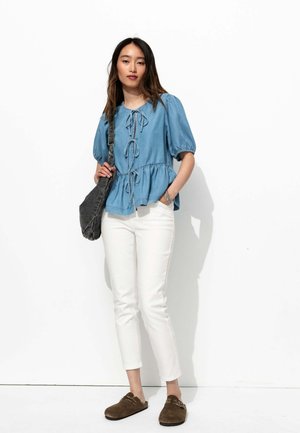 Blouse en denim bleu clair avec des manches bouffantes et un nœud à l'avant, associée à un pantalon blanc ajusté et des chaussures brunes à enfiler. Sac à bandoulière gris.