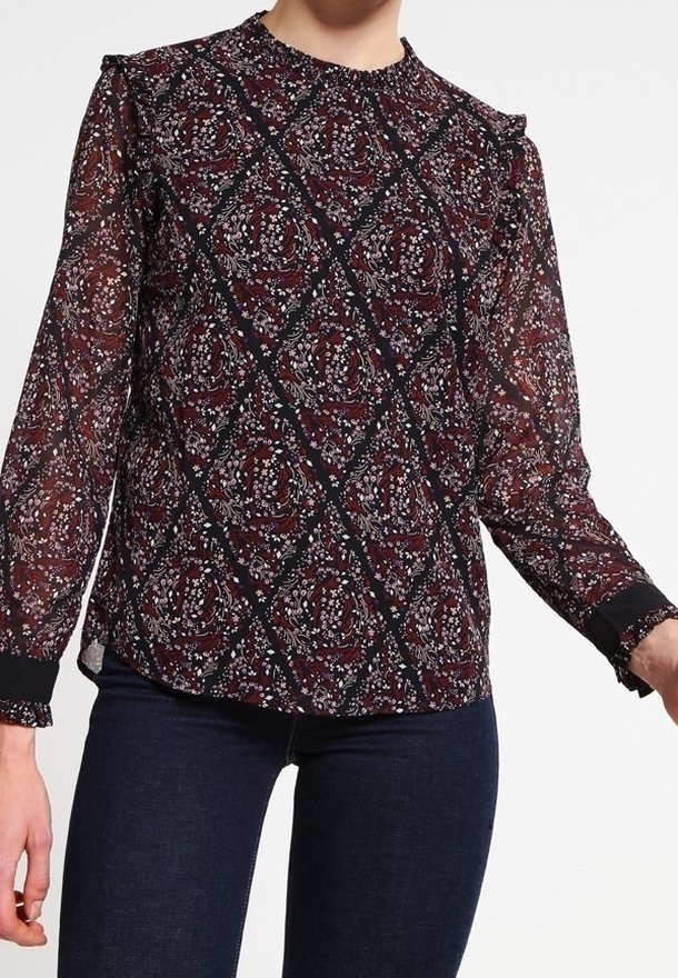 Femme portant une blouse à manches longues à motif floral avec des épaules à volants et un jean foncé sur fond blanc.