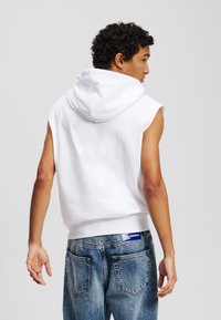Hoodie bianco senza maniche con cappuccio regolabile, abbinato a jeans di denim blu chiaro con tasche posteriori visibili e branding sulla vita.