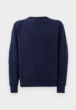 Svetrov navy modre barve s krojem na vratu in dolgimi rokavi ter rebrastimi manšetami in obrobo na enobarvni bež podlagi.