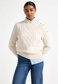 Kremfarvet strikket sweater med diamantmønstre, høj krave og knapdetaljer på skulderen. Kombineret med en blåstribet skjorte.