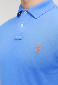 Polo di colore blu chiaro con due bottoni e piccolo logo ricamato di un giocatore di polo arancione sul lato sinistro del petto.