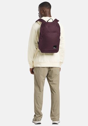 Bordeauxfarbener Rucksack mit glatter Textur, verstellbaren Trägern, Fronttasche und Markenlogo auf einem schwarzen Quadrat. Über einem hellen Hoodie getragen.
