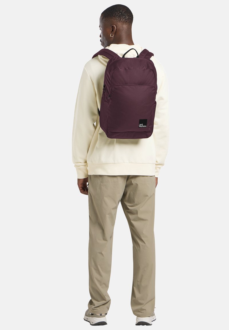 Bordeaux rugzak met een gladde textuur, verstelbare banden, een voorvak en het merkteken op een zwart vierkant. Draagt over een lichtgekleurde hoodie.