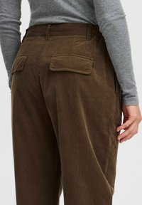 Brown corduroy pants featuring a high waist, two rear pockets with flaps, and a textured finish. Gray long-sleeve shirt partially visible.