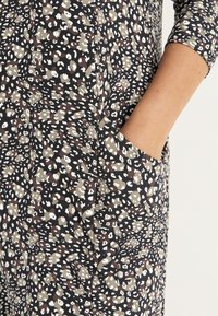 Schwarzes Kleid mit einem gesprenkelten Muster in Beige und Weiß, mit Taschen und Dreiviertelärmeln, aus einem weichen, strukturierten Stoff.