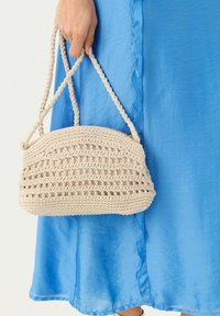 Borsa a tracolla beige all'uncinetto con forma arrotondata e manici intrecciati, caratterizzata da motivi a lavorazione aperta e una texture liscia, tenuta contro un vestito blu.
