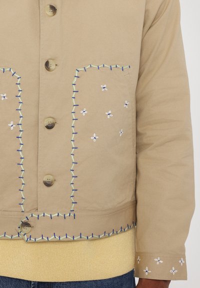 Beige button-up jacket featuring contrasting blue and green stitching, floral embroidery on pockets, and ribbed yellow knit hem and cuffs.