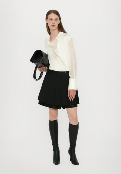 White silk shirt with a relaxed fit, paired with a black pleated skirt and knee-high black leather boots. Model holds a black handbag.