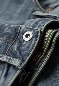 Vaqueros de mezclilla en azul oscuro con una textura suave. Presenta costuras amarillas, un botón de metal con el logotipo "Superdry" y una etiqueta de prenda.