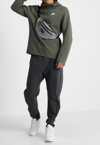 Grön Nike-tröja med långa ärmar kombinerad med mörkgråa mjukisbyxor och vita sneakers. En grå axelväska bärs i midjehöjd.