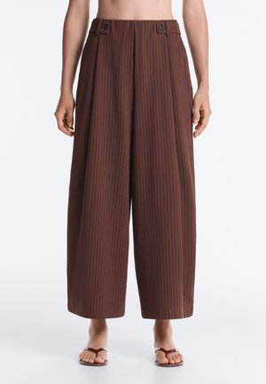 Pantalon large marron à fines rayures avec pattes de taille boutonnées, porté avec des sandales plates marron pieds nus.