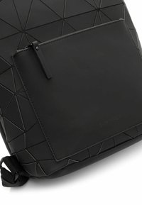 Czarny plecak wykonany z teksturowanego, geometrycznego materiału. Posiada płaską kieszeń z przodu oraz gładką czarną powierzchnię z detalem logo.