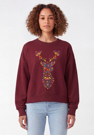 Femme portant un sweat-shirt bordeaux avec un motif coloré de tête de cerf floral, associé à un jean bleu clair, debout sur un fond blanc.