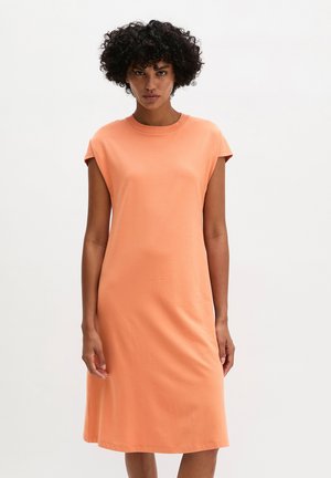 Frau mit lockigem Haar, die ein ärmelloses, knielanges pfirsichfarbenes Kleid trägt und vor einem schlichten hellen Hintergrund steht.