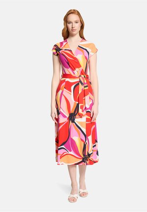 Vrouw draagt een kleurrijke wikkeljurk met grote bloemenpatronen in rood, roze, oranje en zwart, gecombineerd met witte sandalen met open tenen.