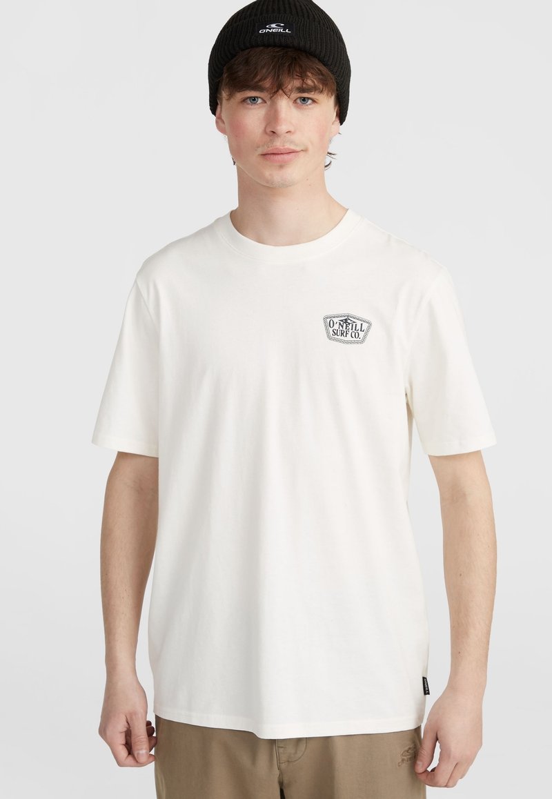 Balts kokvilnas krekls ar īsām piedurknēm un mazu melnu logotipu uz krūtīm. Valkāts ar haki biksēm un melnu beanie cepuri.