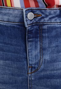 Lähikuva sinisistä farkuista, joiden vyötärö on varustettu metallisella napilla, jossa on oranssia ompeleita, ja monivärinen raidallinen paita on tungettu sisälle.