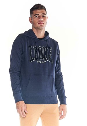 Un homme porte un sweat-shirt à capuche bleu marine avec l’inscription « LEONE 1947 » et un pantalon beige, sur fond blanc.
