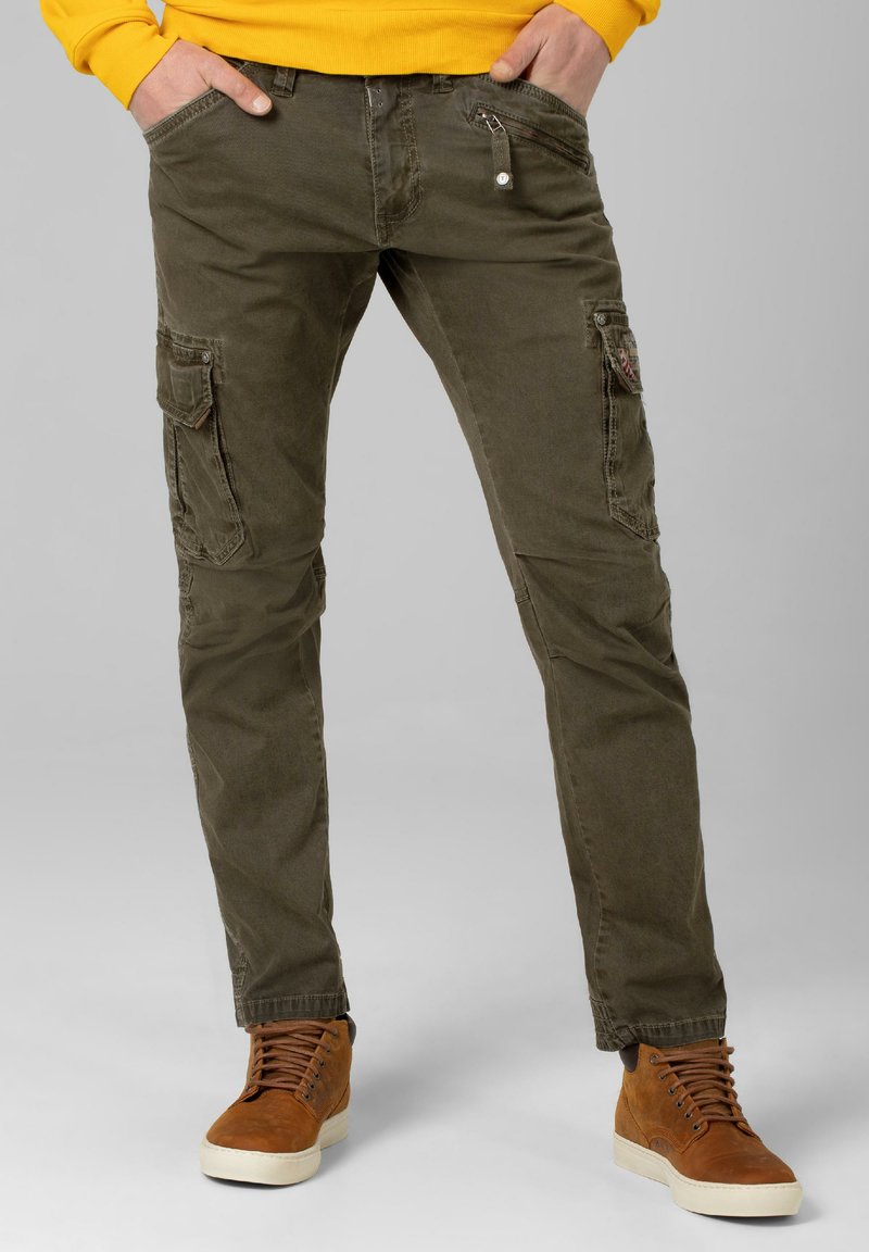 Olivgrüne Cargo-Hosen aus weicher Baumwolle mit mehreren Taschen, Reiß- und Knopfdetails, kombiniert mit braunen High-Top-Sneakern.