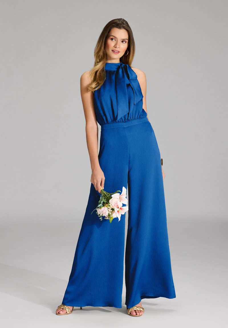 Completo blu con collo all'americana, vita arricciata e pantaloni a gamba larga. Tessuto strutturato con un accento annodato al colletto.