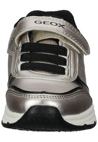 Zapatilla deportiva plateada y negra con tres cordones elásticos negros, una correa de velcro y un cuello acolchado. Presenta materiales suaves y acentos texturizados.