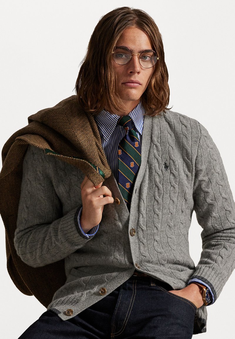 Grijze kabelgebreide cardigan met houten knopen, gelaagd over een blauw-wit gestreept shirt, gecombineerd met een patroonstropdas en een bruine herringbone jas.