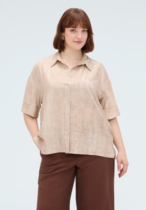 Frau mit kurzen braunen Haaren, die ein beiges, locker sitzendes Hemd mit Knöpfen und braune Hosen trägt, steht mit einer Hand in der Tasche vor hellem Hintergrund.