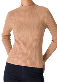 Femme portant un pull beige côtelé à manches longues et un pantalon bleu foncé, debout avec les mains derrière le dos.