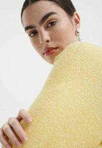 Svijetlo žuti pleteni pulover, meke teksture, sa zaobljenim oblikom. Krupni plan prikazuje facialne osobine i elegantnu frizuru, ruku koja drži pulover.