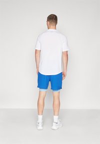 Camisa atlética de manga corta blanca, pantalones cortos azules superpuestos sobre pantalones cortos de compresión azul claro, con zapatos deportivos blancos y detalles de costura visibles.