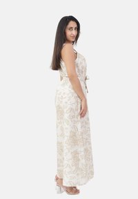 Frau mit langen, dunklen Haaren, die ein weißes, ärmelloses Kleid mit Blumenmuster und transparente High Heels trägt, seitlich stehend und in die Kamera schauend.